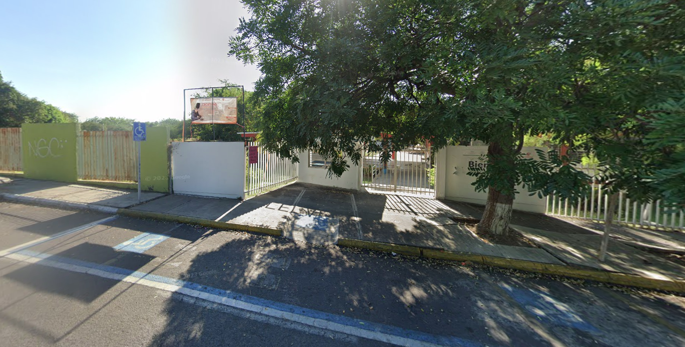 Fotografía en la que se muestra la fachada del plantel, conformada por muros de color verde intercalados con rejas blancas. En medio se encuentra una rampa que da acceso a las instalaciones y al lado derecho de la rampa hay un gran árbol. 